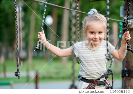 Cute girl enjoying activity in a climbing adventure park. Girl goes on hinged trail. Cute girl enjoying activity in a climbing adventure park. Girl goes on hinged trail. 114056353