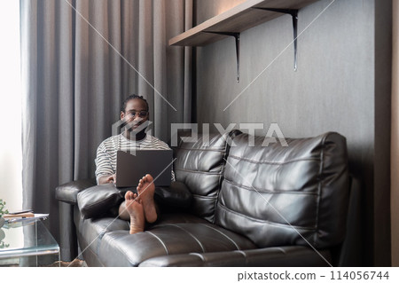Young African American man holding laptop relaxing sitting on sofa working remote at home Young African American man holding laptop relaxing sitting on sofa working remote at home 114056744