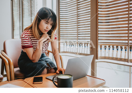 Intently focused freelancer in a striped shirt working on a project on her laptop at a wooden desk by a window. Intently focused freelancer in a striped shirt working on a project on her laptop at a wooden desk by a window. 114058112