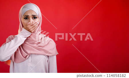 Omg embarrassment. Shame fear. Keep quiet. Overwhelmed frightened sad woman in hijab covering mouth with hand isolated on red empty space background. 114058182