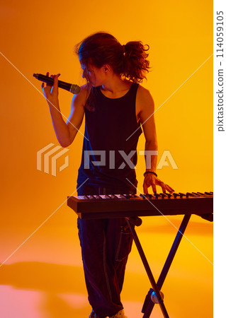 Young man making soulful performance, playing piano and singing against yellow background in neon light 114059105