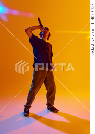 Young emotional guy in casual clothes standing on stage, reading rap against yellow background in neon light 114059190
