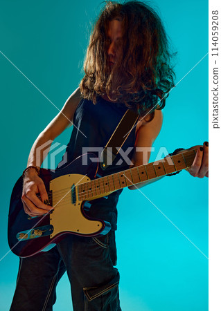 Young man, musician with long hair emotionally and energetically playing guitar in stage against blue, cyan background in neon light Young man, musician with long hair emotionally and energetically playing guitar in stage against blue, cyan background in neon light 114059208