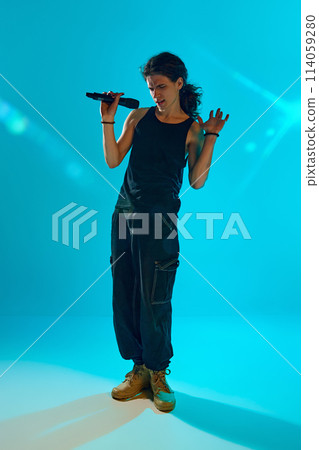 Young man, reading rap, singing on stage, making energetic performance against blue, cyan background in neon light 114059280