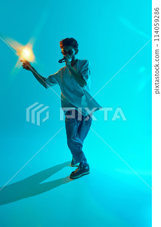With determined expression, young musician delivers powerful rap performance against blue, cyan background in neon light With determined expression, young musician delivers powerful rap performance against blue, cyan background in neon light 114059286