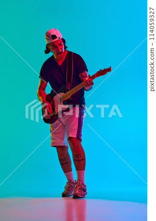 Young man, musician playing electric guitar with intensity and focus against blue, cyan background in neon light 114059327