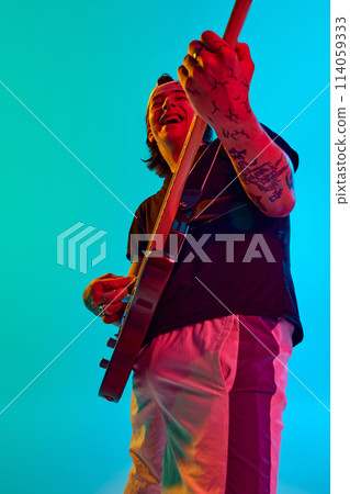 Young emotional man, musician, playing electric guitar, captivating audience with energy of music against blue, cyan background in neon light Young emotional man, musician, playing electric guitar, captivating audience with energy of music against blue, cyan background in neon light 114059333