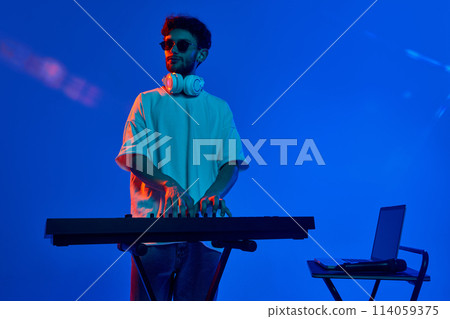 Stylish young man in casual clothes, sunglasses and headphones, making dynamic beats with dj mixer against blue background in neon light Stylish young man in casual clothes, sunglasses and headphones, making dynamic beats with dj mixer against blue background in neon light 114059375
