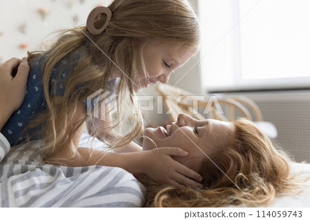 Cheerful little daughter and loving mother lying together on bed Cheerful little daughter and loving mother lying together on bed 114059743