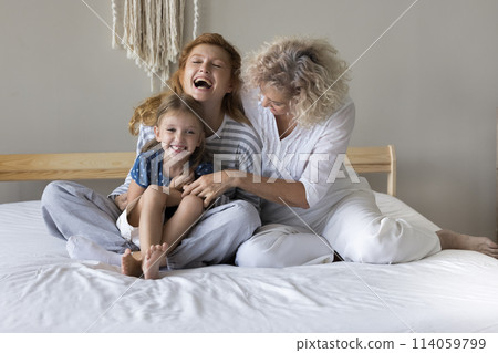 Two generations of women tickling little girl seated on bed 114059799