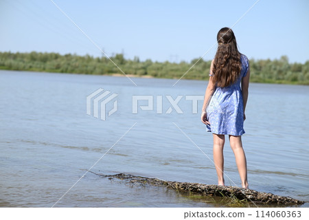 A young girl with long dark hair stands on the bank of the river. 114060363