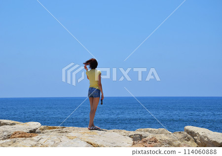 Portrait of a slender woman 30-35 years old in blue shorts and a yellow T-shirt, standing on the seashore and looking into the distance. The concept of rest, travel. People in the back. Portrait of a slender woman 30-35 years old in blue shorts and a yellow T-shirt, standing on the seashore and looking into the distance. The concept of rest, travel. People in the back. 114060888
