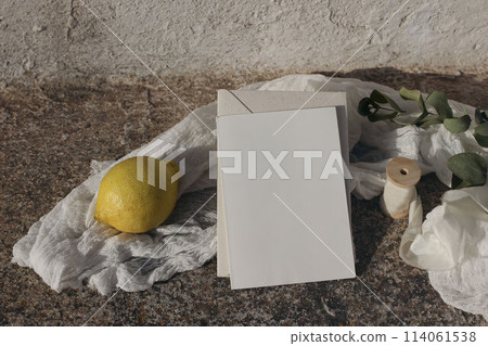 Summer wedding stationery. Blank greeting card invitation mockup in sunlight. Eucalyptus branch, muslin table runner. Fresh lemon fruit. Blurred white shabby old textured wall background. Top view 114061538
