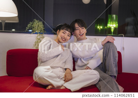Smiling young man and woman sitting side by side on a sofa Smiling young man and woman sitting side by side on a sofa 114062651