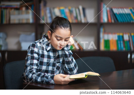beautiful schoolgirl sitting in the library and reading a book 114063640