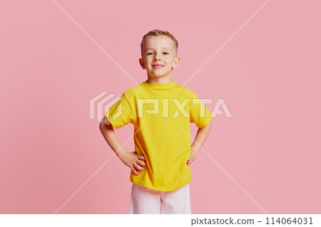 Cute little boy in casual clothes posing holds hands on hips and smiling looks at camera against pink studio background. 114064031