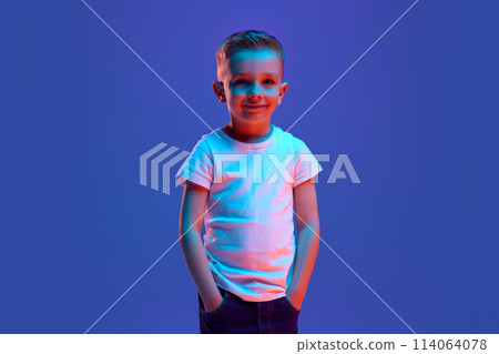 Cheerful smiling little boy dressed casual attire posing looking at camera in mixed neon light against blue background. 114064078