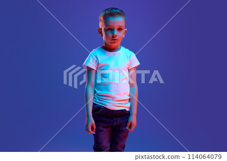 Little boy, student in casual clothe posing looking at camera with calm expression in mixed neon light against blue background. Human emotions. 114064079