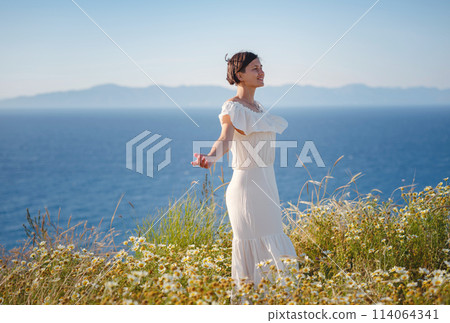 Beautiful Asian young woman in white dress outdoor in flower field under Rhodes city and above sea during sunset. embracing fresh air and engaging in outdoor activities. strong wind - element of 114064341