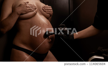 The master evens out an instant tan with a brush on the skin of a pregnant woman. Close-up of the tummy. The master evens out an instant tan with a brush on the skin of a pregnant woman. Close-up of the tummy. 114065604