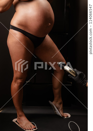 Faceless pregnant woman getting an instant tan. The master applies bronzer with a spray. Vertical photo. Faceless pregnant woman getting an instant tan. The master applies bronzer with a spray. Vertical photo. 114066547
