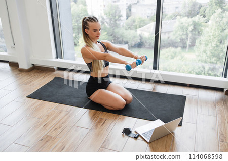 Young woman exercising at home with dumbbells and laptop Young woman exercising at home with dumbbells and laptop 114068598