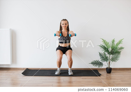 Young Woman Performing Squat Exercise with Dumbbells Indoors Young Woman Performing Squat Exercise with Dumbbells Indoors 114069060