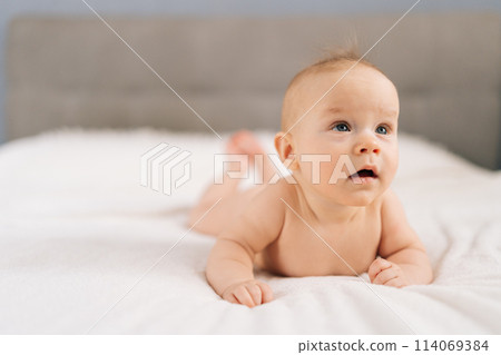 Closeup of cute little baby girl lying on stomach on bed, learning to hold head. Portrait of nice newborn naked baby looking interestingly away with open mouth. Carefree healthy babyhood concept. 114069384