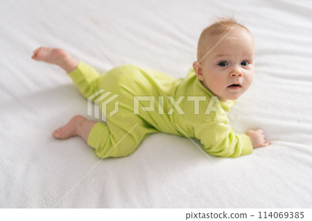 High-angle view of adorable newborn baby girl wearing yellow cotton bodysuit lying on stomach on white sheet. Newborn child relaxing in bed in children nursery, looking at camera. 114069385
