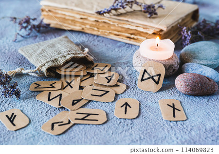 Fortune telling cardboard runes scattered from a bag, a candle, an old book and stones on the table 114069823