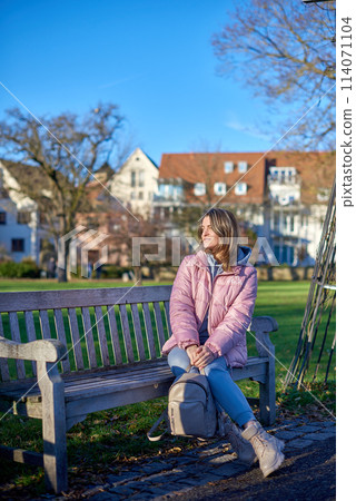 Winter Joy in Bitigheim-Bissingen: Beautiful Girl in Pink Jacket Sitting Amidst Half-Timbered Charm. beautiful girl in a pink winter jacket sitting on a bench in a park, set against the backdrop of 114071104