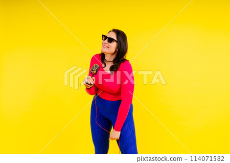 Woman in sport clothes sings into a wired microphone and claps her hands on a yellow background Woman in sport clothes sings into a wired microphone and claps her hands on a yellow background 114071582
