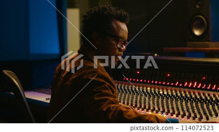 Portrait of african american audio technician uses mixing console with knobs and sliders in control room, editing recordings and adding sound effects. Skilled music producer at mixer. Camera A. Portrait of african american audio technician uses mixing console with knobs and sliders in control room, editing recordings and adding sound effects. Skilled music producer at mixer. Camera A. 114072523