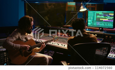Rockstar recording music on his guitar in professional studio, creating new music for his album in control room. Artist composer producing tracks on electro acoustic guitar. Camera B. 114072560