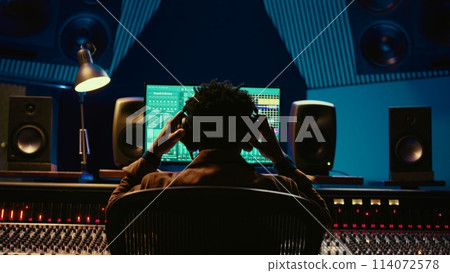 Sound designer editing tracks by adjusting volume levels with panel board knobs and sliders, studio control room. Audio technician mixing and mastering hit song with digital software. Camera B. Sound designer editing tracks by adjusting volume levels with panel board knobs and sliders, studio control room. Audio technician mixing and mastering hit song with digital software. Camera B. 114072578