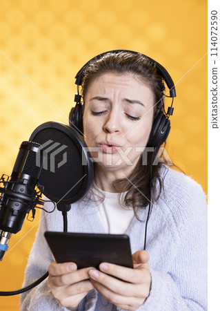 Woman frowns while doing voiceover reading of ebook on ereader to produce audiobook using dramatic acting. Narrator glowers, portraying character, recording novel using digital tablet, studio 114072590