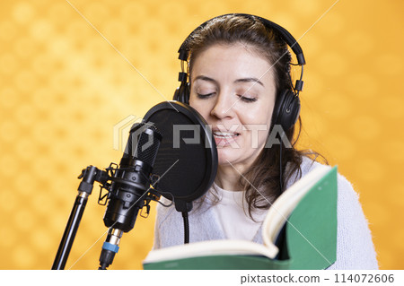 Smiling lady doing voiceover reading of book to produce audiobook. Happy voice actor using storytelling skills to entertain audience while producing digital recording of novel, studio background Smiling lady doing voiceover reading of book to produce audiobook. Happy voice actor using storytelling skills to entertain audience while producing digital recording of novel, studio background 114072606