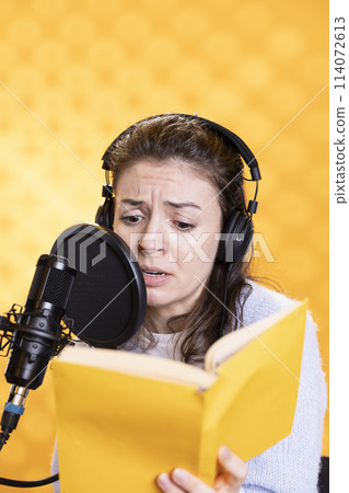 Woman with frown on face wearing headphones reading aloud from book into mic against background. Voice actor recording audiobook, creating engaging content for listeners, glowering for dramatic effect 114072613