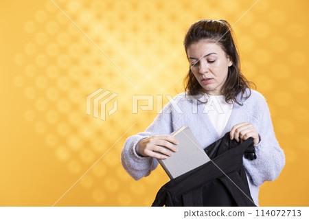 Portrait of woman rummaging through backpack, taking book out, isolated over studio background. Student removing textbook used for educational purposes from school rucksack Portrait of woman rummaging through backpack, taking book out, isolated over studio background. Student removing textbook used for educational purposes from school rucksack 114072713