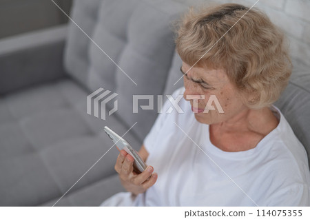 An elderly Caucasian woman suffers from myopia and tries to read a message on a smartphone.  114075355