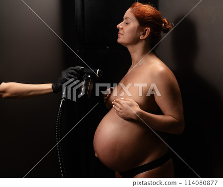 Pregnant woman getting an instant tan. The master applies bronzer with a spray. Pregnant woman getting an instant tan. The master applies bronzer with a spray. 114080877