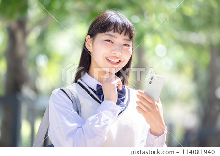A high school girl wearing earphones and looking at her smartphone 114081940