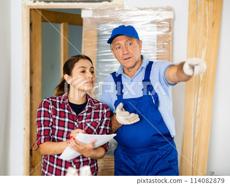 Casual middle-aged repairer in uniform discussing the future construction site with the female in the house Casual middle-aged repairer in uniform discussing the future construction site with the female in the house 114082879