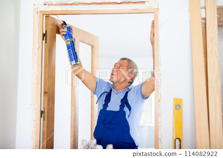 Middle-aged contractor repairman in uniform filling door jambs with polyurethane foam in a house Middle-aged contractor repairman in uniform filling door jambs with polyurethane foam in a house 114082922