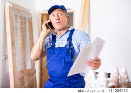 Worried male repairer in uniform talking on a mobile phone while holding a work plan while renovating a house 114084685