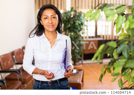Smiling Hispanic businesswoman standing with folder of documents in office Smiling Hispanic businesswoman standing with folder of documents in office 114084714