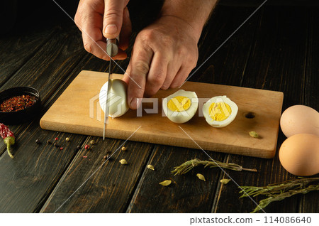 Hands with a knife cut an egg. Sliced eggs on a kitchen board for preparing a diet lunch with aromatic spices. Advertising space Hands with a knife cut an egg. Sliced eggs on a kitchen board for preparing a diet lunch with aromatic spices. Advertising space 114086640