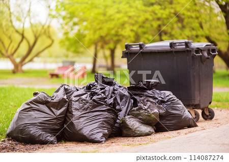 A pile of plastic garbage bags on the side of the road near a plastic container. Garbage bags on the street. Black trash bags. Concept of garbage collection, ecology, environmental cleaning 114087274