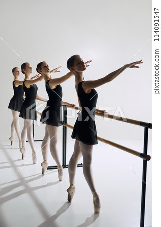 Four ballet dancers, elegant teen girls standing at barre in black costumes and pointe shoes, practicing against grey studio background 114091547