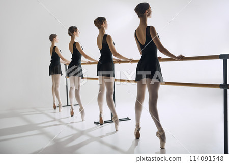 Four ballet dancers, elegant teen girls standing at barre in black costumes and pointe shoes, practicing against grey studio background 114091548
